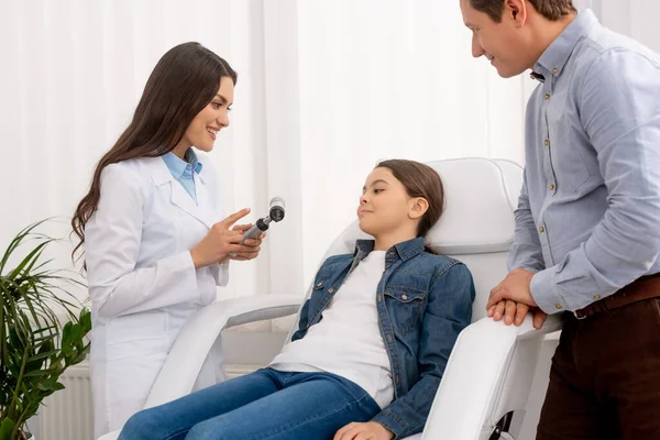 Médico sorrindo ent apontando com o dedo para o otoscópio perto de criança sorridente sentado na cadeira médica e seu pai de pé nas proximidades — Fotografia de Stock