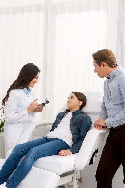Отоларинголог показывает отоскоп улыбающемуся ребенку, сидящему в медицинском кресле, и ее отцу, стоящему рядом — стоковое фото
