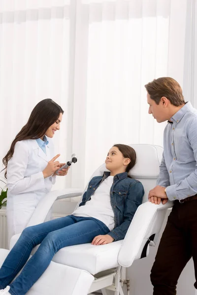 Otolaryngologist showing otoscope to smiling kid sitting in medical chair and her father standing nearby — Stock Photo