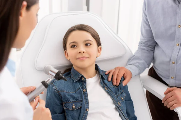 Обрезанный вид отца, трогающего плечо дочери, сидящей в медицинском кресле рядом с отоларингологом с помощью отоскопа — стоковое фото