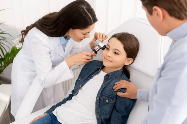 Vista ritagliata del padre toccare spalla di figlia seduta sulla sedia medica mentre otorinolaringoiatra esaminando l'orecchio con otoscopio — Foto stock