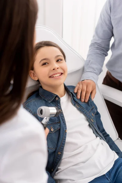 Vue recadrée du père touchant l'épaule de sa fille assise sur une chaise médicale près d'un médecin tenant un otoscope — Photo de stock