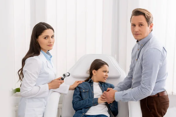 Father holding hands of worried daughter sitting in medical chair near ent physician with otoscope — Stock Photo