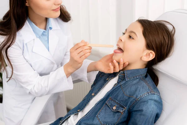 Vista parcial del otorrinolaringólogo examinando la garganta del niño con depresor de la lengua - foto de stock