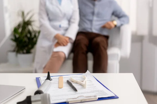 Selective focus of desk with ent diagnostic equipment on clipboard, and otolaryngologist with patient sitting on background — Stock Photo