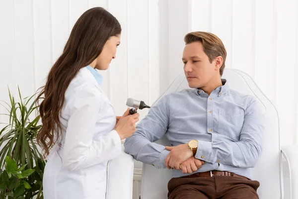 Grazioso medico in possesso di otoscopio vicino bel paziente seduto sulla sedia medica — Foto stock