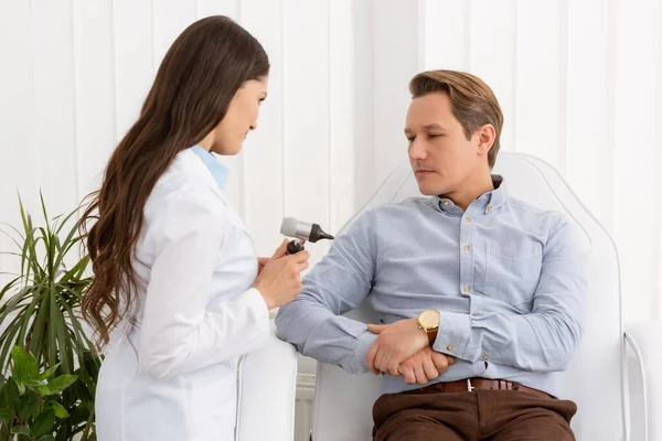 Bastante médico sosteniendo otoscopio cerca hermoso paciente sentado en silla médica - foto de stock