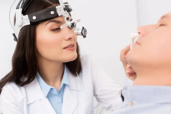 Beautiful, attentive otolaryngologist in ent headlight examining nose of man with nasal speculum — Stock Photo