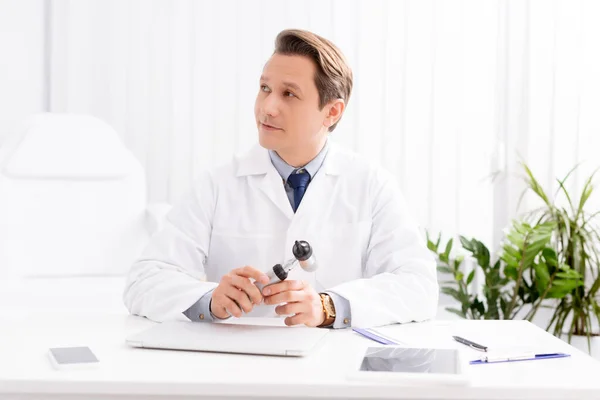 Beau médecin tenant l'otoscope et regardant loin tout en étant assis sur le lieu de travail — Photo de stock