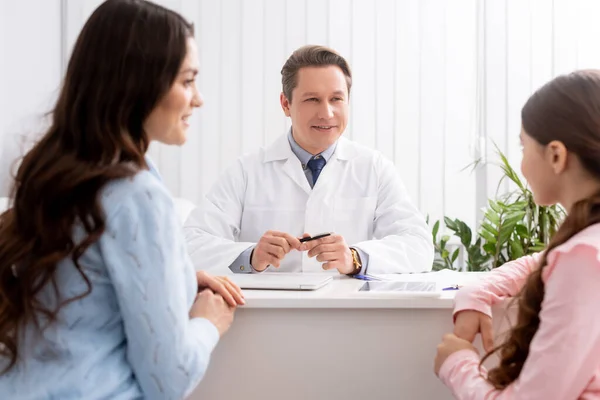 Enfoque selectivo de la madre y la hija sentadas cerca del médico sonriente ent durante la consulta - foto de stock