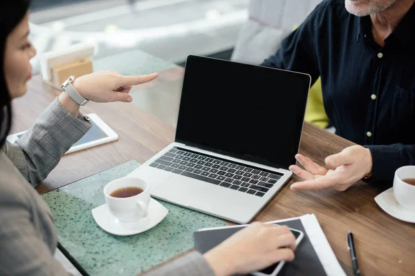 Обрезанный вид деловой женщины, указывающей пальцем на ноутбук и беседующей с бизнесменом во время деловой встречи — стоковое фото