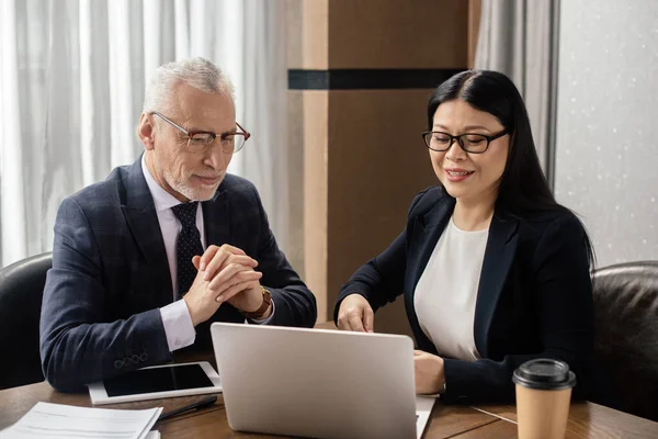 Uomo d'affari e sorridente donna d'affari asiatica guardando il computer portatile durante la riunione di lavoro — Foto stock