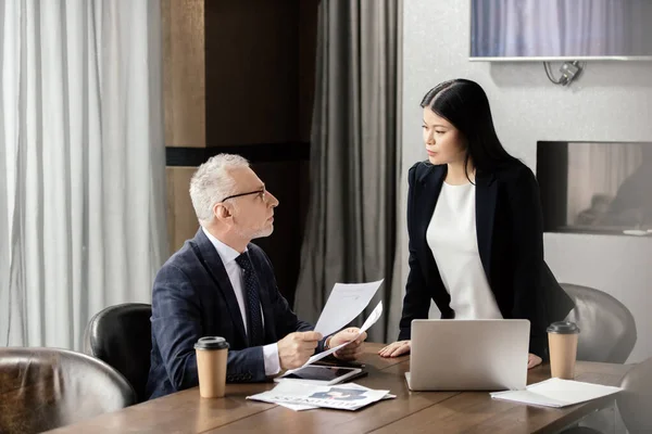 Бизнесмен, держащий бумаги и беседующий с азиатской бизнес-леди во время деловой встречи — стоковое фото