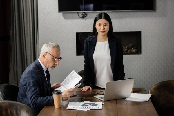 Homme d'affaires utilisant un smartphone et une femme d'affaires asiatique regardant la caméra pendant une réunion d'affaires — Photo de stock