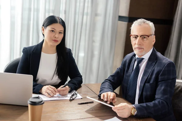 Businessman and asian businesswoman looking at camera and sitting at table — Stock Photo