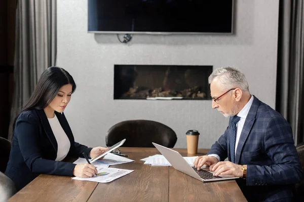 Vista laterale di uomo d'affari utilizzando laptop e donna d'affari asiatica che fa scartoffie durante la riunione di lavoro — Foto stock