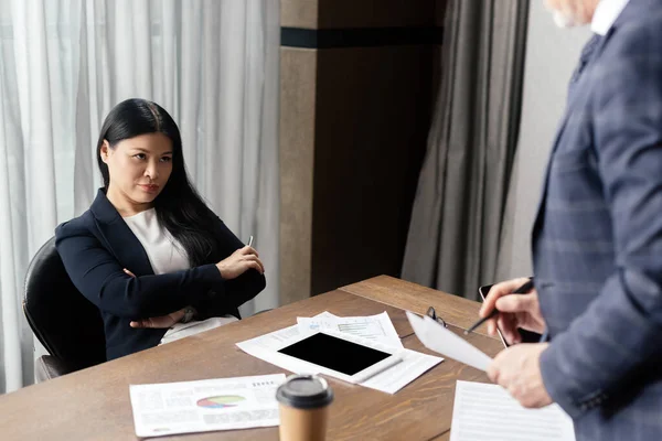 Cropped view of businessman and offended asian businesswoman with crossed arms during business meeting — Stock Photo