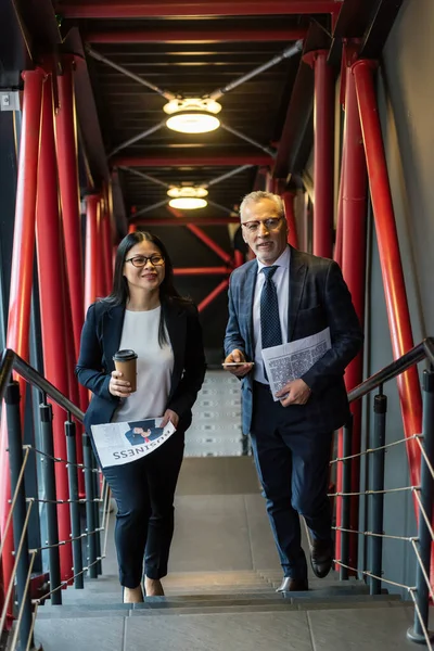 Lächelnder Geschäftsmann und asiatische Geschäftsfrau mit Zeitungen, die in die Kamera schauen — Stockfoto