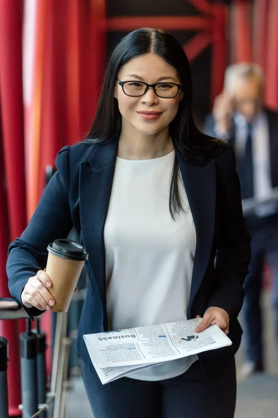 Lächelnde asiatische Geschäftsfrau mit Pappbecher und Zeitung blickt in die Kamera — Stockfoto