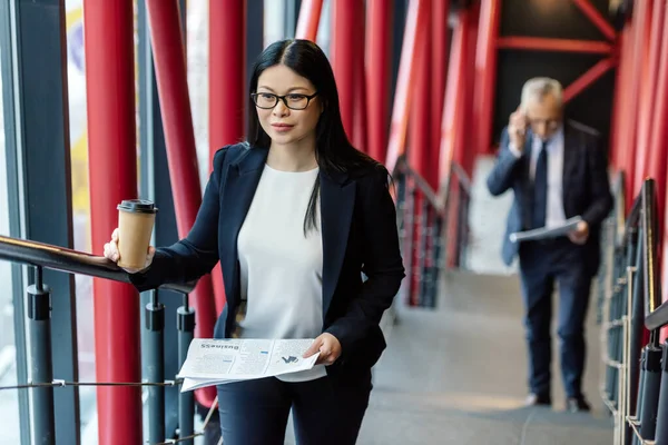 Selective focus of smiling asian businesswoman with paper cup and newspaper looking away — Stock Photo
