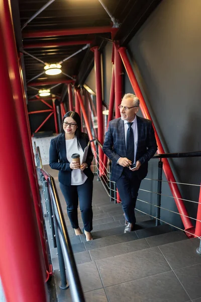 Hochwinkelaufnahme eines lächelnden Geschäftsmannes und einer asiatischen Geschäftsfrau, die auf Treppen reden und gehen — Stockfoto