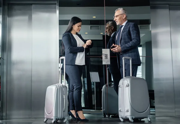 Lächelnder Geschäftsmann und asiatische Geschäftsfrau mit Reisetaschen, die in der Nähe von Aufzügen stehen — Stockfoto
