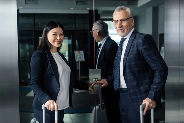 Sonriente hombre de negocios y asiático mujer de negocios con bolsas de viaje pulsando botón de ascensor - foto de stock