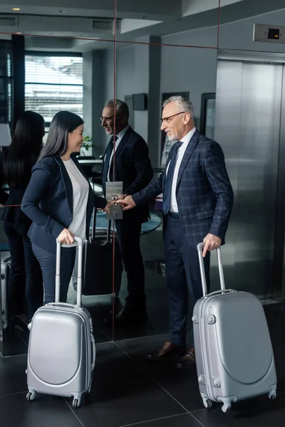 Smiling businessman and asian businesswoman with travel bags pushing button of elevator — Stock Photo