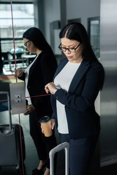 Asiatische Geschäftsfrau mit Pappbecher schaut auf Armbanduhr und steht in der Nähe von Fahrstuhl — Stockfoto