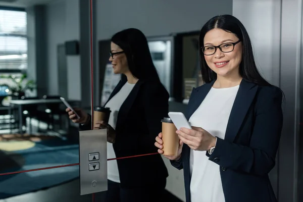 Smiling asian businesswoman holding paper cup, smartphone and looking at camera — Stock Photo