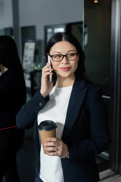 Smiling asian businesswoman holding paper cup and talking on smartphone — Stock Photo