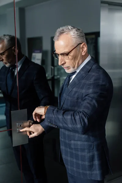 Hombre de negocios en traje mirando el reloj de pulsera y pulsando el botón del ascensor - foto de stock