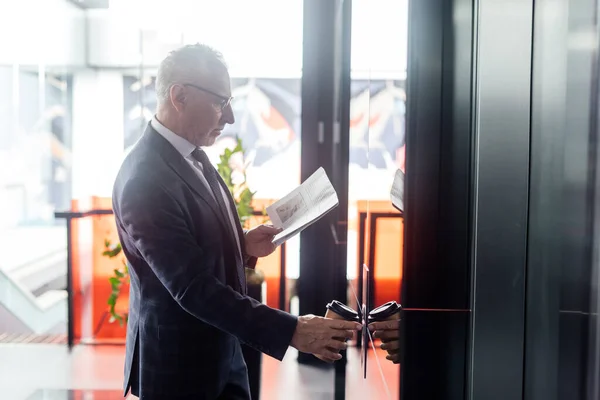 Side view of businessman with newspaper and paper cup pushing button of elevator — Stock Photo