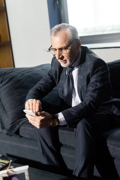 Homem de negócios de terno sentado no sofá e usando smartphone no hotel — Fotografia de Stock