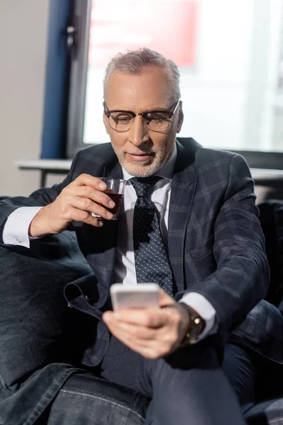 Selective focus of businessman using smartphone and holding glass — Stock Photo