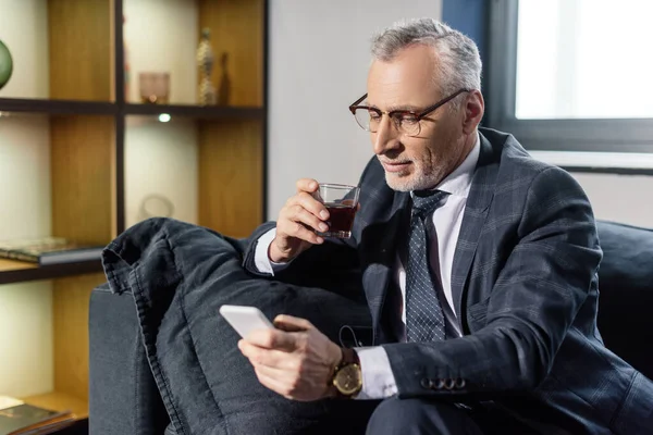 Selective focus of smiling businessman using smartphone and holding glass — Stock Photo