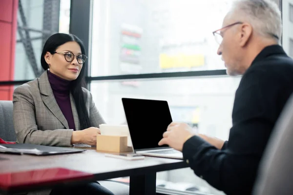 Selective focus of smiling asian businesswoman talking with businessman in cafe — Stock Photo