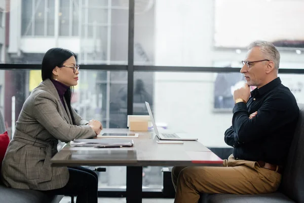 Vista laterale di uomo d'affari e donna d'affari asiatica seduto a tavola e parlando in caffè — Foto stock