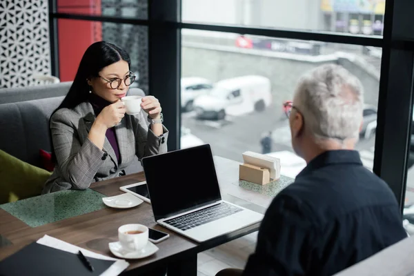 Enfoque selectivo de la mujer de negocios asiática beber té y sentarse a la mesa con el hombre de negocios - foto de stock