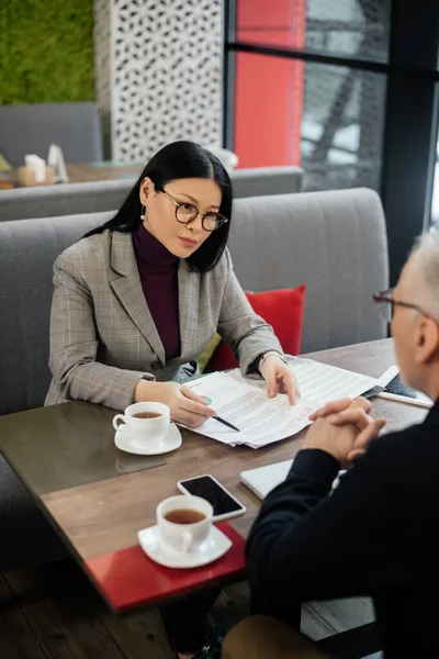 Enfoque selectivo de la mujer de negocios asiática hablando con el hombre de negocios y apuntando a los papeles en la cafetería - foto de stock
