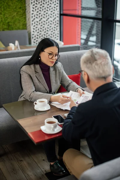 Enfoque selectivo de la mujer de negocios asiática hablando con el hombre de negocios y la celebración de papeles en la cafetería - foto de stock