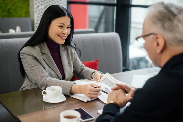 Enfoque selectivo de sonriente mujer de negocios asiática hablando con hombre de negocios y apuntando a los papeles en la cafetería - foto de stock