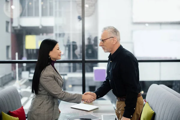 Side view of businessman and smiling asian businesswoman shaking hands in cafe — Stock Photo