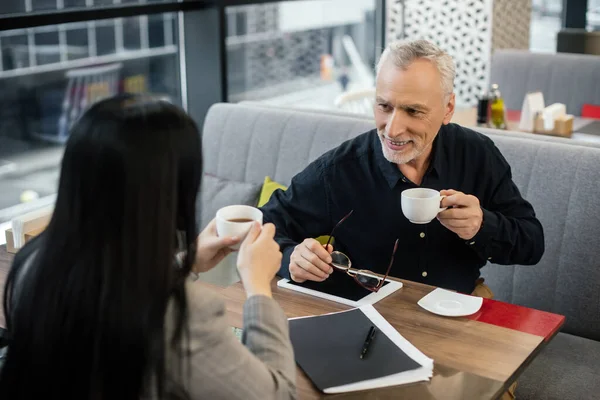 Focalizzazione selettiva di un uomo d'affari sorridente che tiene la tazza e parla con una donna d'affari nel caffè — Foto stock