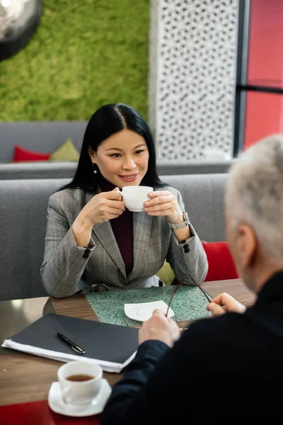 Enfoque selectivo de sonreír mujer de negocios asiática hablando con el hombre de negocios y la celebración de la taza en la cafetería - foto de stock