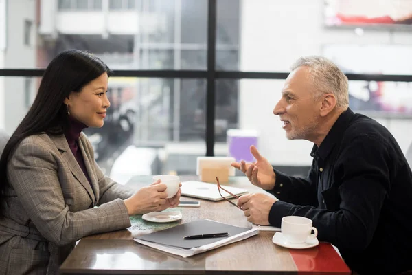 Vista lateral de hombre de negocios y sonriente mujer de negocios asiática hablando en cafetería - foto de stock