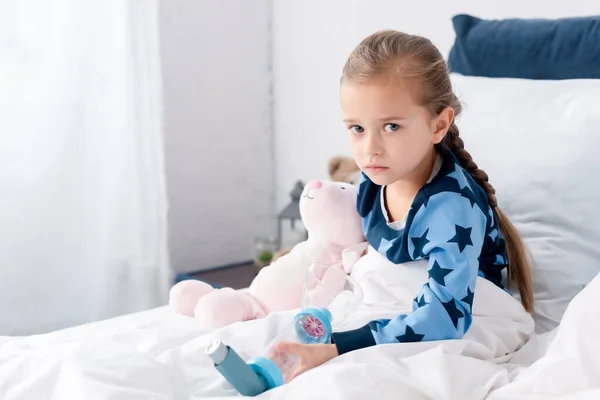 Niño enfermo sosteniendo inhalador con espaciador y mirando a la cámara en el dormitorio - foto de stock