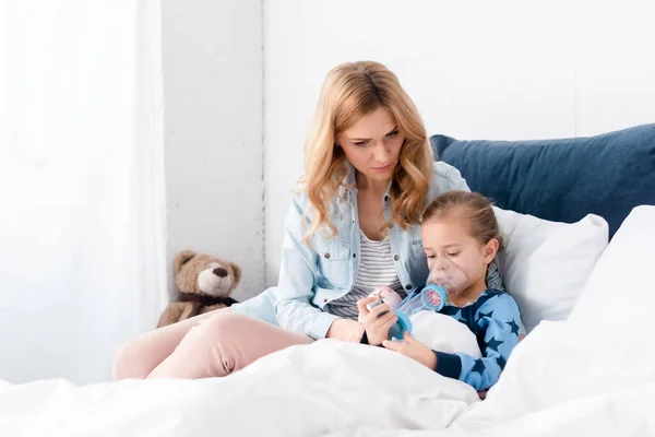 Madre cariñosa sentada con hija enferma usando inhalador con espaciador en el dormitorio - foto de stock