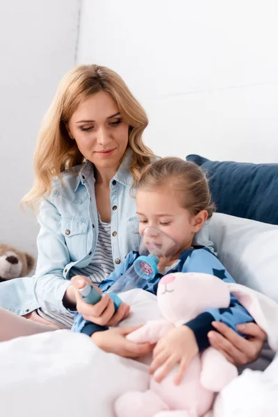 Caring mother sitting near sick daughter using inhaler with spacer and holding soft toy — Stock Photo