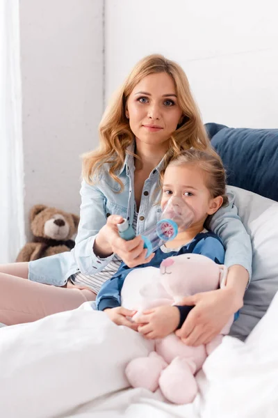 Atractiva madre sentada cerca de hija enferma usando inhalador con espaciador - foto de stock